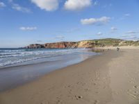 two people walking on the beach under a partly cloudy sky, and an ocean with a large cliff in the background