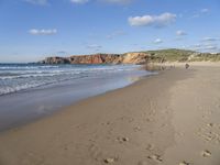 two people walking on the beach under a partly cloudy sky, and an ocean with a large cliff in the background