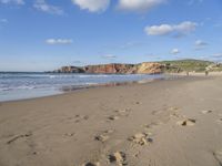 two people walking on the beach under a partly cloudy sky, and an ocean with a large cliff in the background