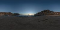 two people are sitting on the sand by the water at sunset overlooking a rock formation