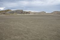 the beach is brown with many sand and some waves crashing along it with a large rock outcropping to the side