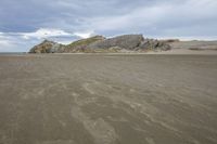 the beach is brown with many sand and some waves crashing along it with a large rock outcropping to the side