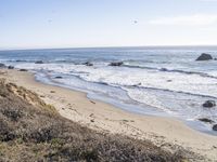 beach scene with water and sand near grassy hills and rocks near ocean shore with rocks on shoreline