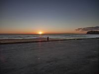 two people are sitting on an empty bench in front of the ocean at sunset with a dog in the foreground