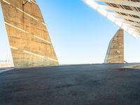 a skateboarder riding around an empty parking space under a large structure with tall triangular pillars