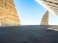 a skateboarder riding around an empty parking space under a large structure with tall triangular pillars