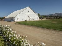 there is a white barn with flowers in the foreground and mountains in the distance