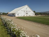 there is a white barn with flowers in the foreground and mountains in the distance