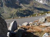 a mountain road is lined with boulders and rocks next to a mountain range with tall, grassy hills in the distance