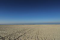 people walk on sand at a beach, near the ocean in the distance is a sky with few cloudless, and blue skies