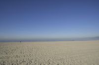 people walk on sand at a beach, near the ocean in the distance is a sky with few cloudless, and blue skies