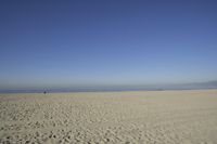 people walk on sand at a beach, near the ocean in the distance is a sky with few cloudless, and blue skies