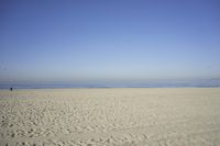 people walk on sand at a beach, near the ocean in the distance is a sky with few cloudless, and blue skies