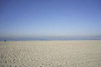 people walk on sand at a beach, near the ocean in the distance is a sky with few cloudless, and blue skies