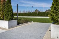 a view of a park with some stone walkways and bushes in the background with flags flying behind a statue