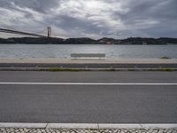 a bench is sitting on a street by the water near a bridge over a body of water