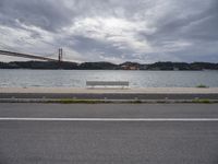 a bench is sitting on a street by the water near a bridge over a body of water