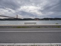 a bench is sitting on a street by the water near a bridge over a body of water