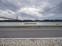 a bench is sitting on a street by the water near a bridge over a body of water