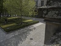 two benches sitting outside on the side of a sidewalk next to a building with a brick walkway that looks out over a formal garden