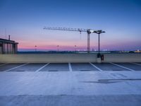 cranes in the air against a dusk sky at an industrial area with parking spaces and city buildings