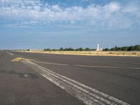 the empty runway has a single airplane in it and a bright blue sky with clouds