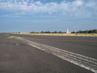 the empty runway has a single airplane in it and a bright blue sky with clouds