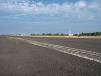 the empty runway has a single airplane in it and a bright blue sky with clouds