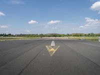 a runway with a yellow arrow painted on the ground in front of an airport and trees in the distance