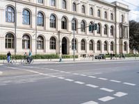 people crossing the street in front of an elegant building and cyclists are passing by on bicycles