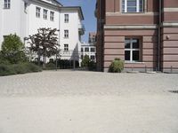 a courtyard has bushes and trees on it in front of buildings in the background and a bench to the left of the driveway