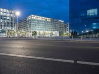 two glass and steel buildings at night near a street with street lights on it and one building with multiple floors