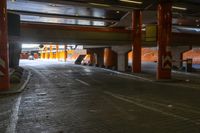 an empty sidewalk beneath a bridge with orange columns and poles at night time with light from overhead