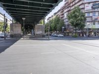 cars and pedestrians pass under a train track in an old part of the city, with trees lining either side