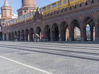 an empty road with large stone walls and towers on it in an urban setting, with buildings lining the streets and trolley tracks passing by