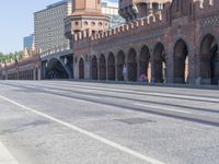 an empty road with large stone walls and towers on it in an urban setting, with buildings lining the streets and trolley tracks passing by