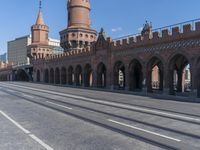 an empty road with large stone walls and towers on it in an urban setting, with buildings lining the streets and trolley tracks passing by