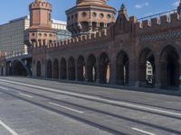 an empty road with large stone walls and towers on it in an urban setting, with buildings lining the streets and trolley tracks passing by