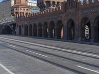 an empty road with large stone walls and towers on it in an urban setting, with buildings lining the streets and trolley tracks passing by