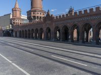 an empty road with large stone walls and towers on it in an urban setting, with buildings lining the streets and trolley tracks passing by