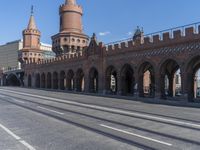 an empty road with large stone walls and towers on it in an urban setting, with buildings lining the streets and trolley tracks passing by