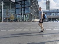 man in shorts and backpack walking across street in front of a large glass building with multiple levels