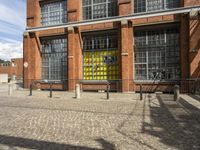 a brick sidewalk lined with a yellow gate in front of a building with windows and bikes parked outside