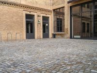 two doors of a building in a courtyard next to a fence and a bench on the side of a brick road