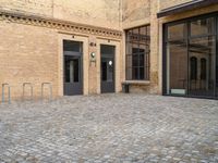 two doors of a building in a courtyard next to a fence and a bench on the side of a brick road