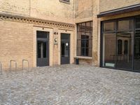 two doors of a building in a courtyard next to a fence and a bench on the side of a brick road