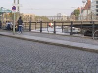 the woman is walking near her bicycle on the sidewalk of a bridge that crosses a canal