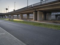 an empty sidewalk near an overpass leading to highway traffic in the evening hours, with a bench under a bridge and traffic light