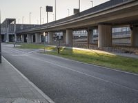 an empty sidewalk near an overpass leading to highway traffic in the evening hours, with a bench under a bridge and traffic light