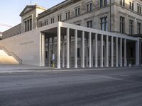 the large, empty sidewalk with multiple columns in front of the building with two signs